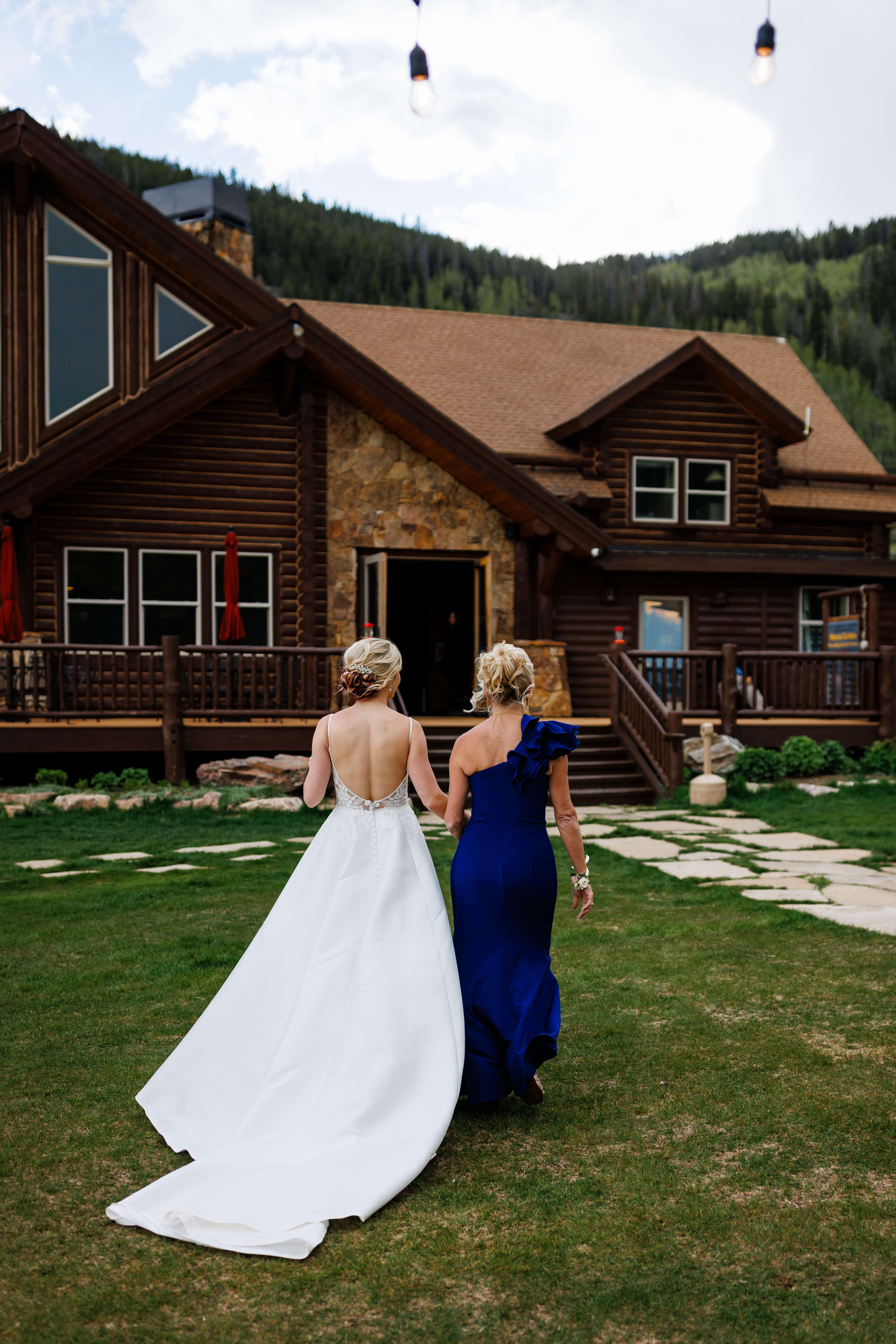 Two women in elegant dresses walking hand in hand towards a rustic wooden cabin in a lush green setting.