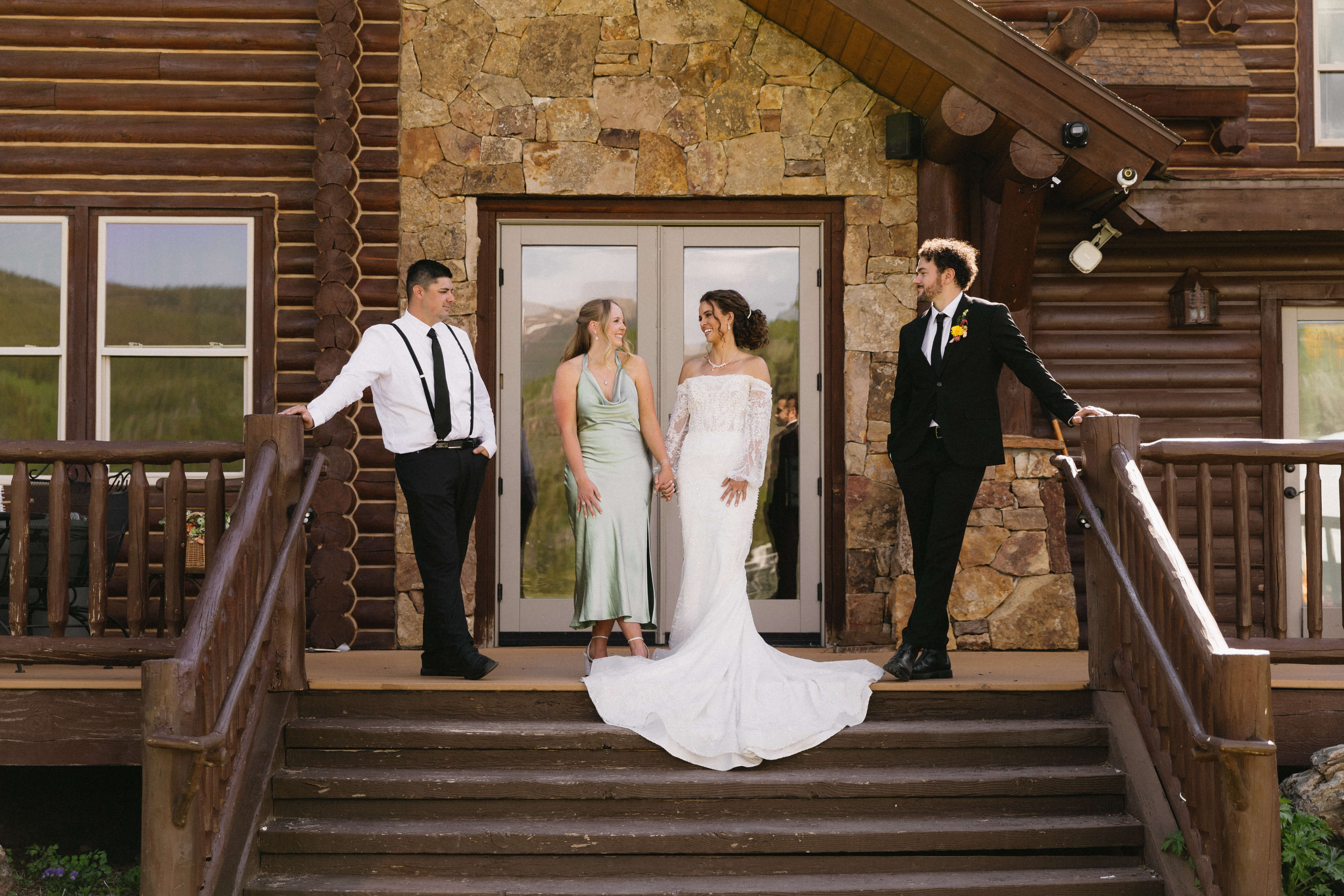 A bride and three people in formal attire posing for photos on a log cabin's steps.