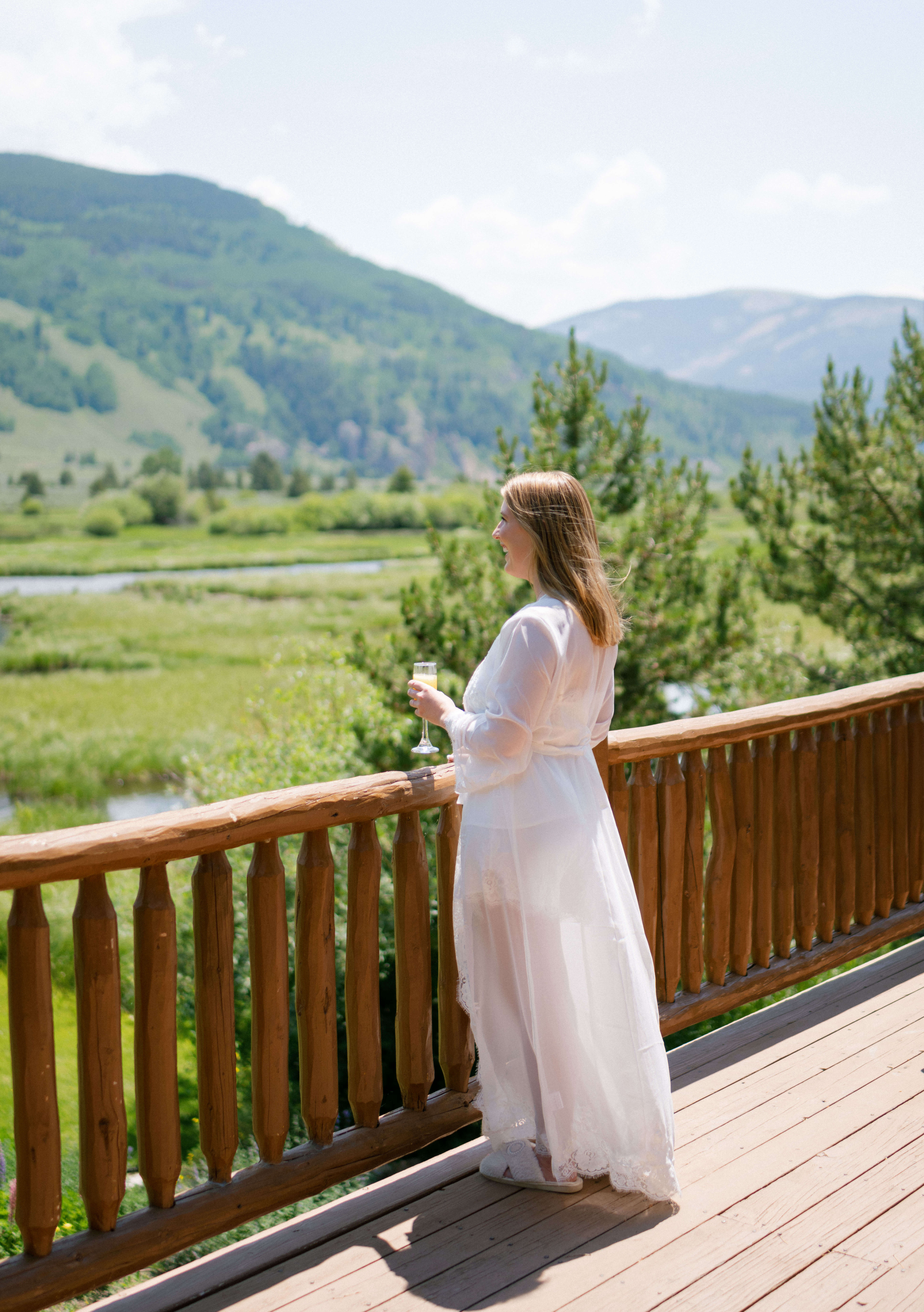 A woman in a white wedding dress holding a glass, standing on a wooden deck overlooking a mountainous landscape.