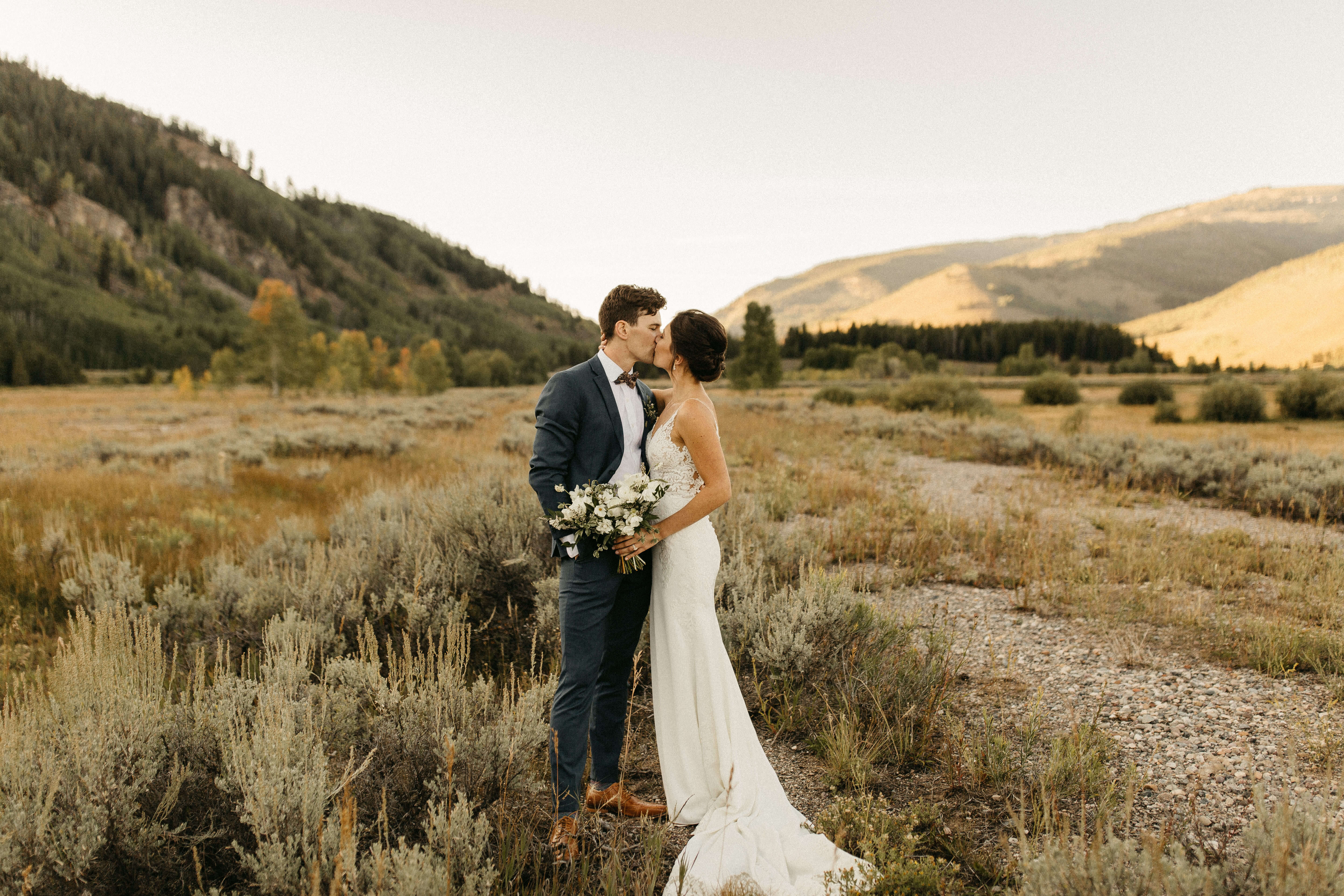 A couple in wedding attire sharing a kiss in a scenic, mountainous field.