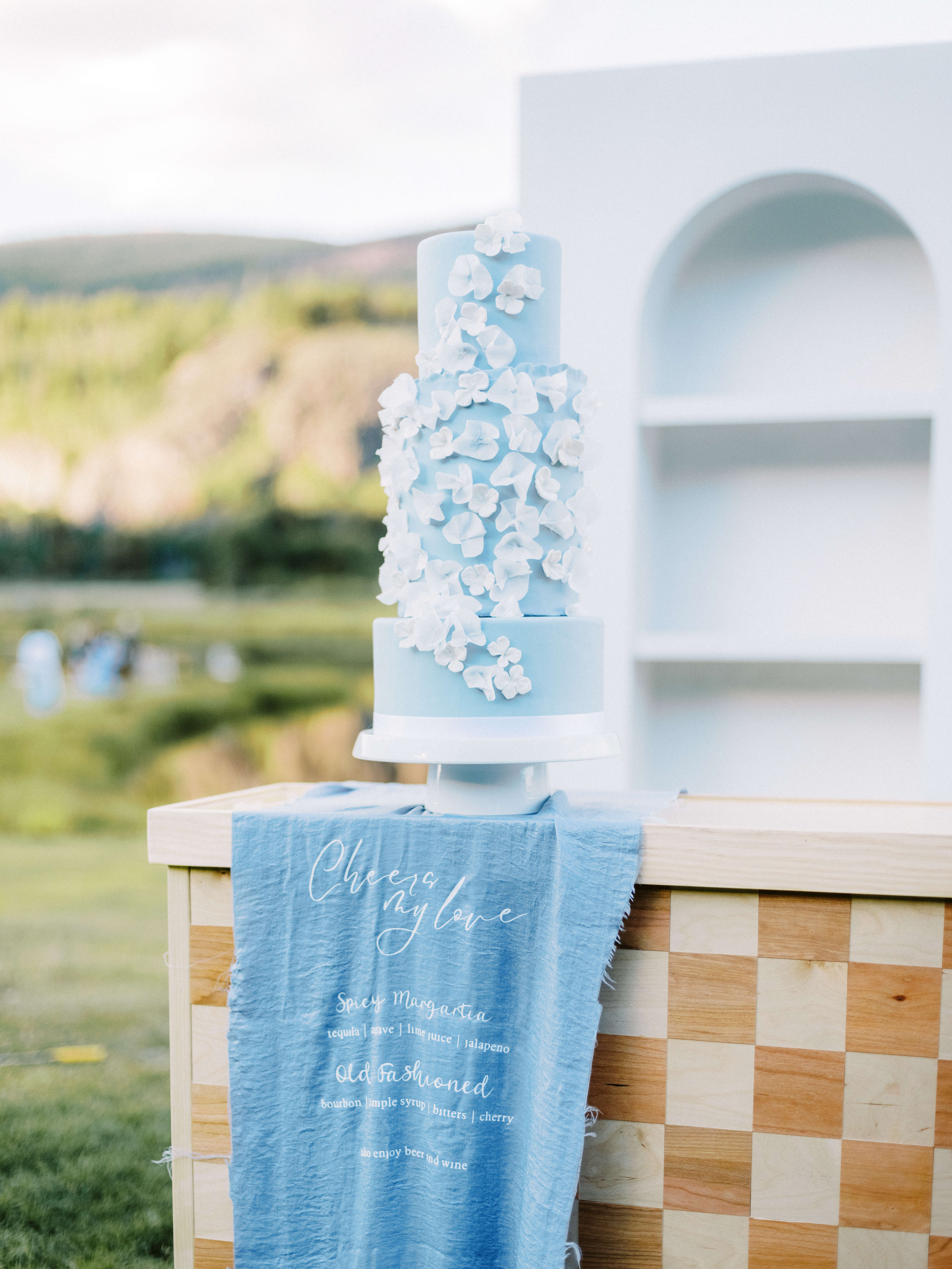 A tall blue wedding cake with white flowers, on a wooden table with a blue cloth, set outdoors.